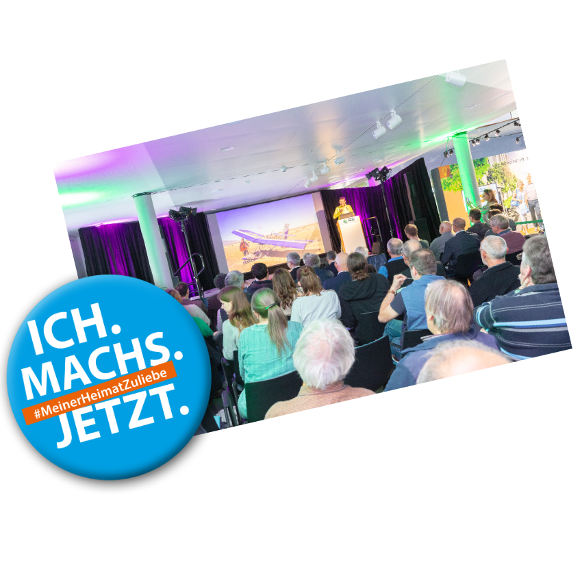 Badge mit Text: "Ich machs jetzt #MeinerHeimatZuliebe" auf einem Bild in dem im Vordergrund ein Publikum zu sehen ist, dahinter steht eine Person am Rednerpult vor einer Leinwand auf der ein Flugzeug zu sehen ist.